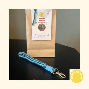 May include: A macrame wristlet keyring kit in a brown paper bag. The kit includes coloured cords and a keyring. The finished wristlet is blue and green with a gold-coloured clasp. The bag has text that reads "Crafting Calm."