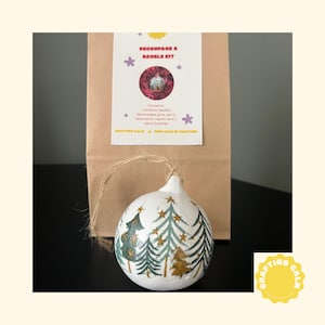 Puede incluir: Una bola de cerámica blanca con un diseño de bosque, parte de un kit de manualidades. La bola presenta árboles y estrellas verdes y doradas. El kit incluye una bola de cerámica, pegamento para decoupage, una servilleta decorativa y pinceles. El kit está en una bolsa de papel marrón.