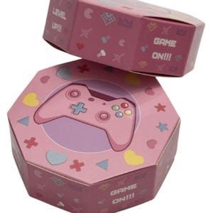 May include: Pink octagonal gift boxes with a video game theme. The top box reads "LEVEL UP!!!" and the bottom box features a pink game controller. The boxes are decorated with hearts, stars, and other game-related icons. The text "GAME ON!!!" is also visible.