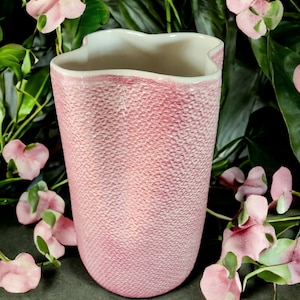 May include: A pink ceramic vase with a textured, pebbled exterior surface and a wavy, scalloped rim. The vase has a tapered cylindrical shape that widens slightly toward the top, featuring a smooth, off-white interior finish visible at the opening.