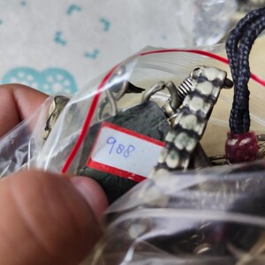 Puede incluir: Una colecci&oacute;n de piezas de joyer&iacute;a met&aacute;lica dentro de una bolsa de pl&aacute;stico transparente, que incluye un colgante de piedra verde oscuro con una etiqueta blanca rectangular con el n&uacute;mero 988 escrito en azul, junto a eslabones plateados y cord&oacute;n negro.