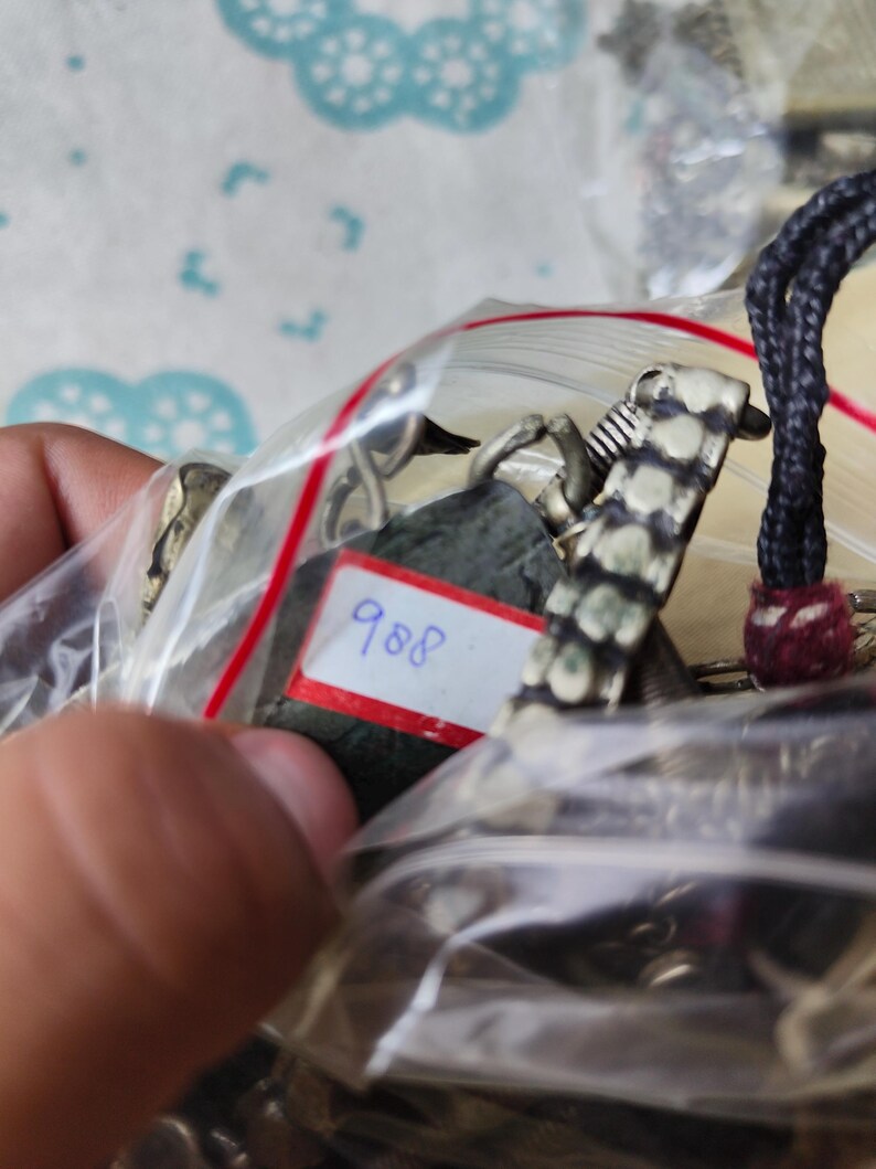 Puede incluir: Una colecci&oacute;n de piezas de joyer&iacute;a met&aacute;lica dentro de una bolsa de pl&aacute;stico transparente, que incluye un colgante de piedra verde oscuro con una etiqueta blanca rectangular con el n&uacute;mero 988 escrito en azul, junto a eslabones plateados y cord&oacute;n negro.