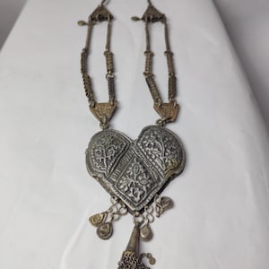 Puede incluir: Un collar de metal plateado con un gran colgante en forma de coraz&oacute;n con grabados florales intrincados. El colgante est&aacute; adornado con dijes colgantes y una peque&ntilde;a borla de diminutos discos met&aacute;licos. La cadena consta de eslabones met&aacute;licos texturizados.