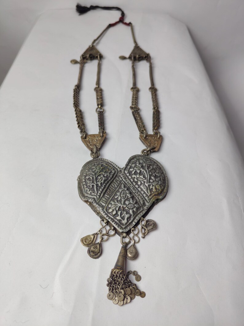 Puede incluir: Un collar de metal plateado con un gran colgante en forma de coraz&oacute;n con grabados florales intrincados. El colgante est&aacute; adornado con dijes colgantes y una peque&ntilde;a borla de diminutos discos met&aacute;licos. La cadena consta de eslabones met&aacute;licos texturizados.