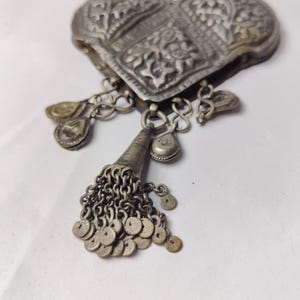 Puede incluir: Un colgante en forma de coraz&oacute;n de tono plateado con intrincados patrones florales en relieve. La pieza incluye varios dijes colgantes peque&ntilde;os y un componente c&oacute;nico central con un racimo de diminutos discos circulares en cadenas.