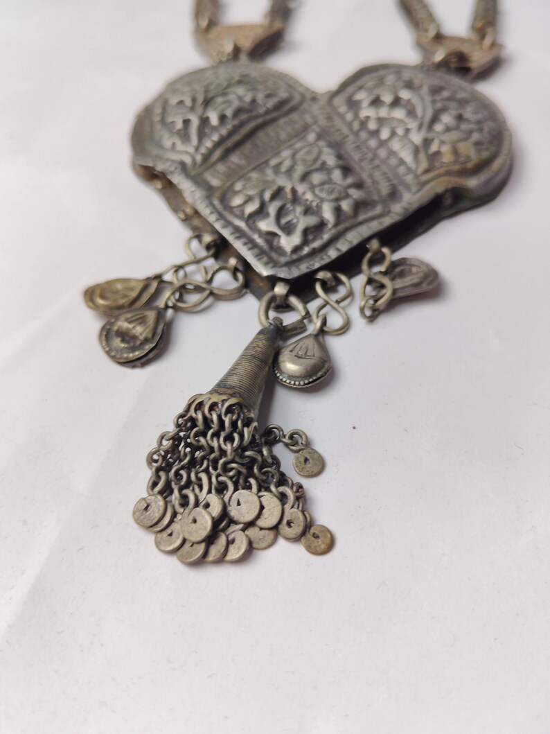 Puede incluir: Un colgante en forma de coraz&oacute;n de tono plateado con intrincados patrones florales en relieve. La pieza incluye varios dijes colgantes peque&ntilde;os y un componente c&oacute;nico central con un racimo de diminutos discos circulares en cadenas.