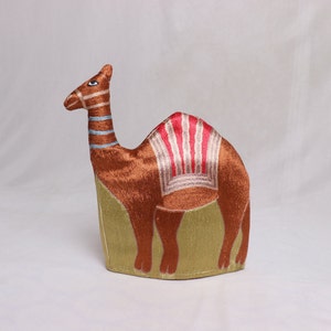 May include: A brown camel plush toy with a red and white striped saddle. The camel is sitting on a green background.