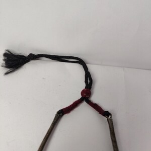 Puede incluir: Un collar que presenta dos tubos met&aacute;licos en espiral conectados por una uni&oacute;n de hilo rojo oscuro. La secci&oacute;n superior consiste en un cord&oacute;n negro con una peque&ntilde;a cuenta roja y un extremo de borla negra deshilachada para el ajuste.