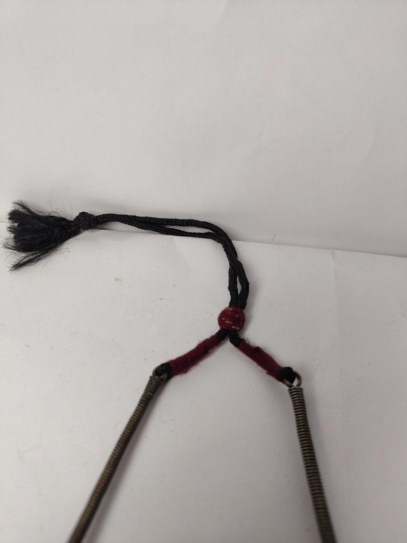 Puede incluir: Un collar que presenta dos tubos met&aacute;licos en espiral conectados por una uni&oacute;n de hilo rojo oscuro. La secci&oacute;n superior consiste en un cord&oacute;n negro con una peque&ntilde;a cuenta roja y un extremo de borla negra deshilachada para el ajuste.