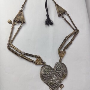 Puede incluir: Un collar de metal plateado con un gran colgante en forma de coraz&oacute;n con grabados florales intrincados. El collar incluye detalles triangulares laterales, eslabones texturizados y peque&ntilde;os dijes colgantes en la parte inferior, terminado con un cord&oacute;n negro.