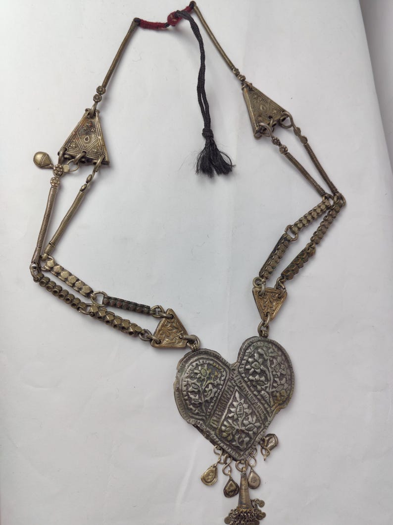 Puede incluir: Un collar de metal plateado con un gran colgante en forma de coraz&oacute;n con grabados florales intrincados. El collar incluye detalles triangulares laterales, eslabones texturizados y peque&ntilde;os dijes colgantes en la parte inferior, terminado con un cord&oacute;n negro.