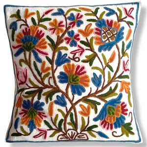 May include: Square white decorative pillow with an embroidered floral design. The pillow features a detailed pattern of colorful flowers in shades of blue, orange, red, and pink, with green leaves and brown stems. The pillow has a blue border.