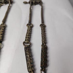 Puede incluir: Un collar de metal plateado con componentes superiores triangulares, segmentos de alambre enrollado y eslabones rectangulares con una superficie texturizada y facetada. La pieza est&aacute; construida con m&uacute;ltiples cadenas y elementos met&aacute;licos interconectados.