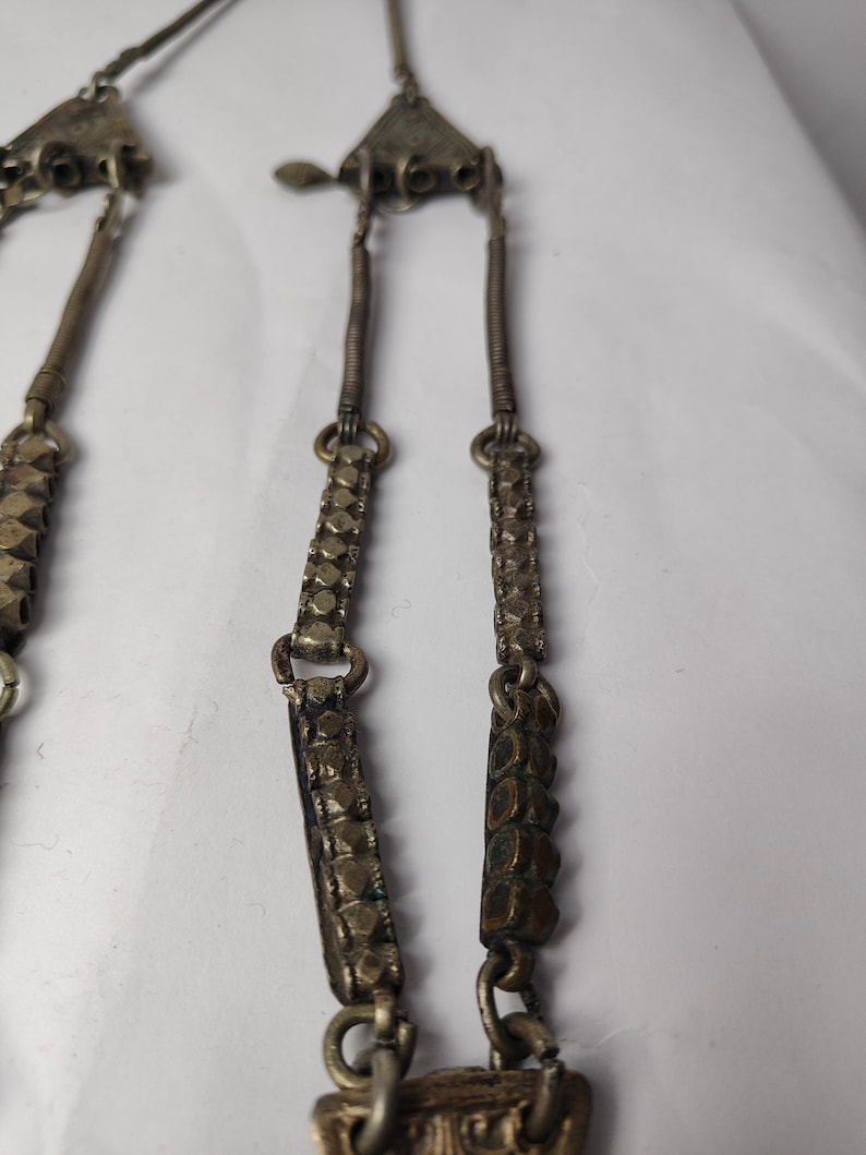 Puede incluir: Un collar de metal plateado con componentes superiores triangulares, segmentos de alambre enrollado y eslabones rectangulares con una superficie texturizada y facetada. La pieza est&aacute; construida con m&uacute;ltiples cadenas y elementos met&aacute;licos interconectados.