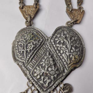 Puede incluir: Un collar con colgante en forma de coraz&oacute;n de tono plateado con patrones florales en relieve y un dise&ntilde;o central en forma de diamante. El colgante cuelga de dos cadenas met&aacute;licas texturizadas unidas por placas triangulares grabadas e incluye peque&ntilde;os dijes.