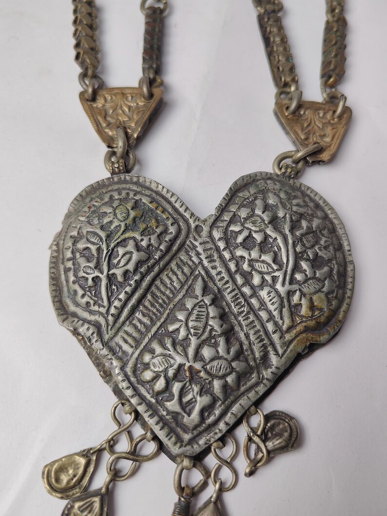 Puede incluir: Un collar con colgante en forma de coraz&oacute;n de tono plateado con patrones florales en relieve y un dise&ntilde;o central en forma de diamante. El colgante cuelga de dos cadenas met&aacute;licas texturizadas unidas por placas triangulares grabadas e incluye peque&ntilde;os dijes.
