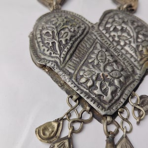 Puede incluir: Un colgante de metal plateado en forma de coraz&oacute;n con patrones florales en relieve y detalles texturizados. El colgante cuelga de una cadena e incluye varios peque&ntilde;os dijes en forma de l&aacute;grima unidos al borde inferior.