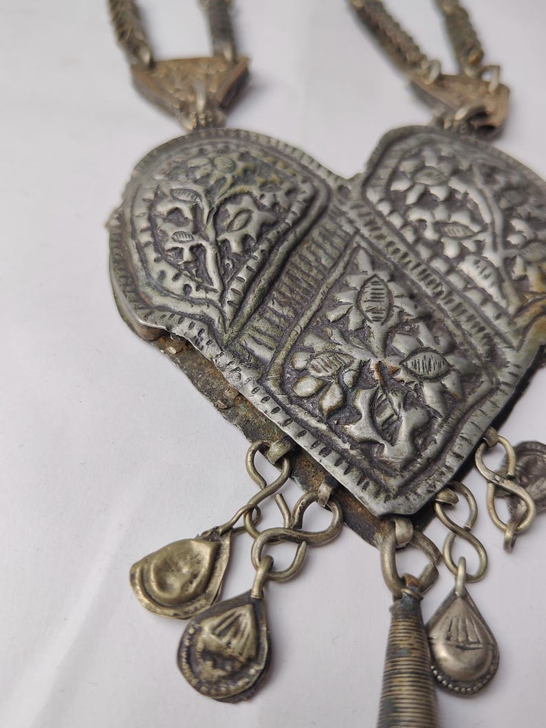 Puede incluir: Un colgante de metal plateado en forma de coraz&oacute;n con patrones florales en relieve y detalles texturizados. El colgante cuelga de una cadena e incluye varios peque&ntilde;os dijes en forma de l&aacute;grima unidos al borde inferior.