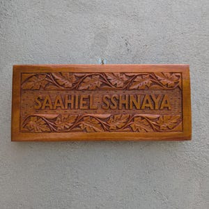 Pode incluir: Uma placa retangular de madeira com o nome "SAAHIEL SSHNAYA" entalhado. A placa é emoldurada com um padrão detalhado de folhas e videiras. A madeira tem um tom castanho quente.