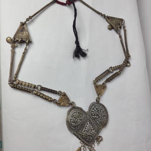 Puede incluir: Un collar met&aacute;lico con un gran colgante en forma de coraz&oacute;n con intrincados grabados florales. La cadena consta de eslabones met&aacute;licos segmentados, placas decorativas triangulares y peque&ntilde;os dijes colgantes. Un cord&oacute;n negro cierra la pieza.