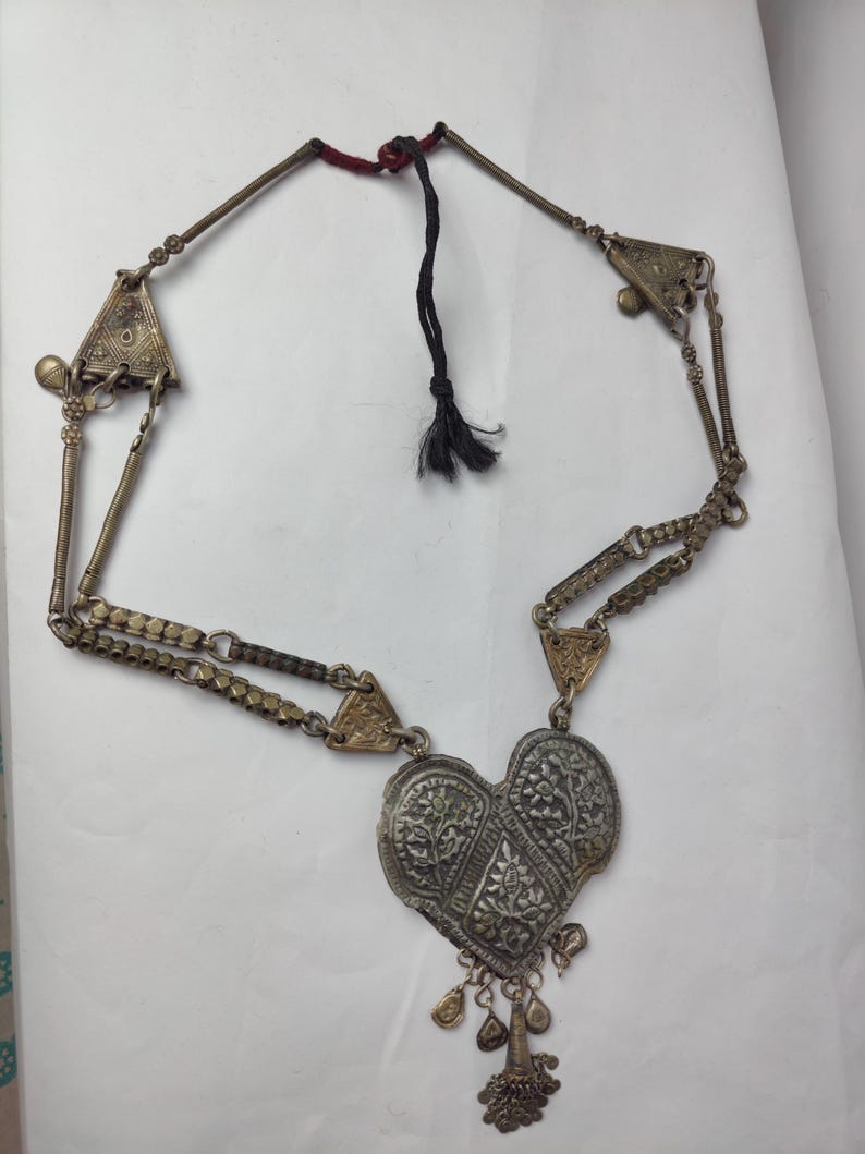 Puede incluir: Un collar met&aacute;lico con un gran colgante en forma de coraz&oacute;n con intrincados grabados florales. La cadena consta de eslabones met&aacute;licos segmentados, placas decorativas triangulares y peque&ntilde;os dijes colgantes. Un cord&oacute;n negro cierra la pieza.