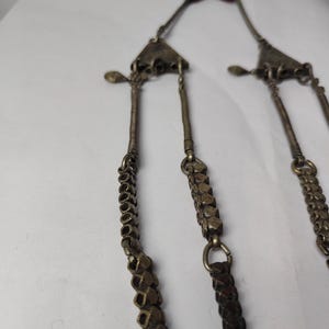 Puede incluir: Un collar met&aacute;lico con dos colgantes triangulares conectados a varias cadenas colgantes. Las cadenas est&aacute;n compuestas por cuentas texturizadas de tono plateado oscuro y segmentos de alambre enrollado, terminando en un cord&oacute;n negro con borla.