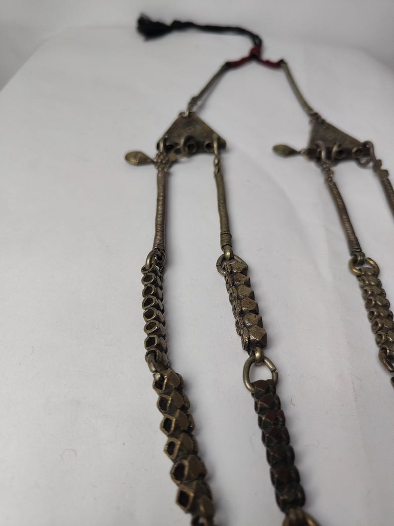 Puede incluir: Un collar met&aacute;lico con dos colgantes triangulares conectados a varias cadenas colgantes. Las cadenas est&aacute;n compuestas por cuentas texturizadas de tono plateado oscuro y segmentos de alambre enrollado, terminando en un cord&oacute;n negro con borla.