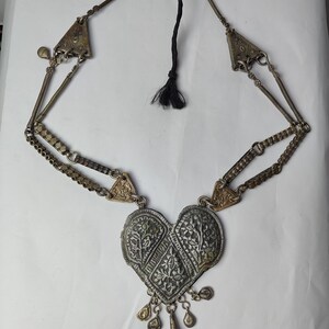 Puede incluir: Un collar de metal plateado con un gran colgante en forma de coraz&oacute;n con intrincados grabados florales. El colgante tiene peque&ntilde;os dijes colgantes en la parte inferior. La cadena consta de eslabones de metal texturizados y detalles triangulares.