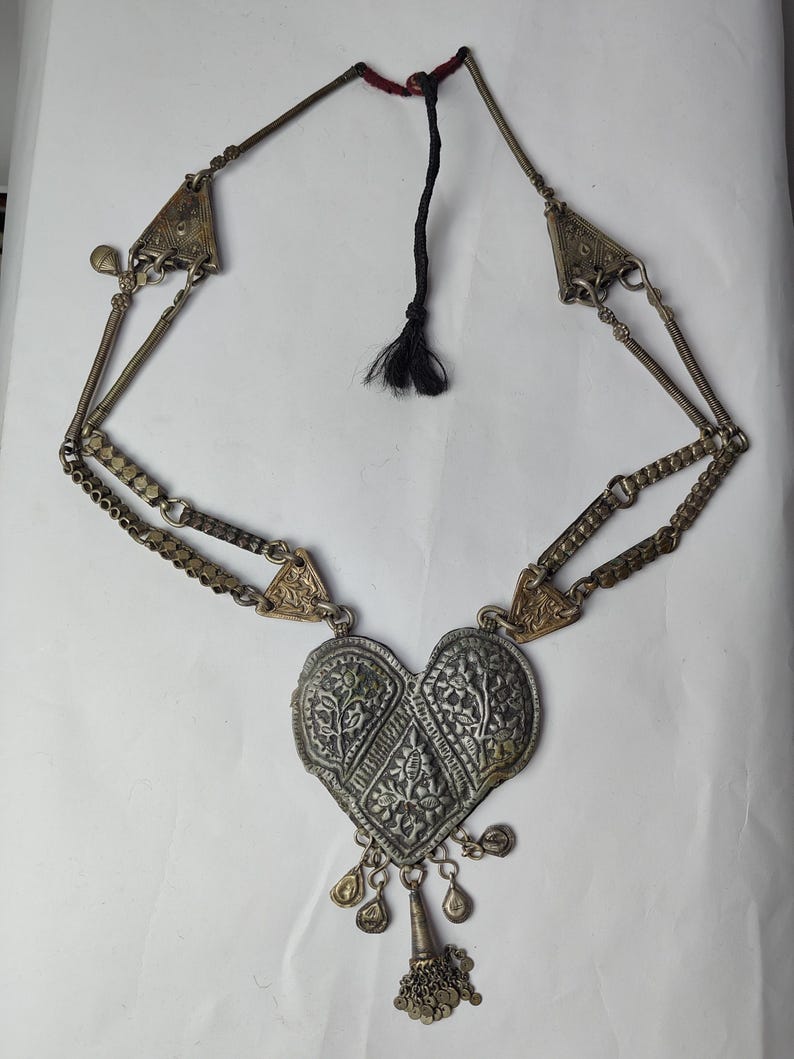 Puede incluir: Un collar de metal plateado con un gran colgante en forma de coraz&oacute;n con intrincados grabados florales. El colgante tiene peque&ntilde;os dijes colgantes en la parte inferior. La cadena consta de eslabones de metal texturizados y detalles triangulares.