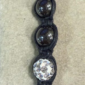 May include: A black beaded bracelet featuring alternating black, glossy beads and sparkling, crystal-covered beads. The bracelet is strung on black cord, creating a striking contrast between the dark beads and the shimmering crystals.