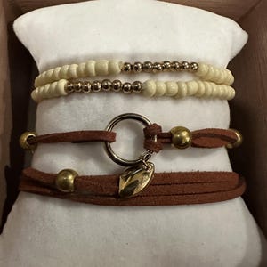 May include: A collection of bracelets displayed on a white cushion. Two bracelets feature cream-colored beads and gold-colored accents. Two brown leather wrap bracelets have gold-colored beads and a gold leaf charm.