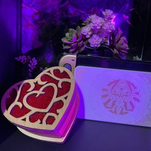 May include: A wooden heart-shaped box with a red glitter interior and a cutout design. The box is sitting on a black surface next to a white card with a gold design. The card has the Triforce symbol from the Legend of Zelda video game series.