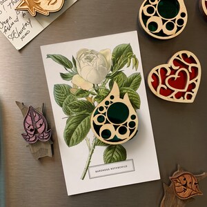 K&ouml;nnte beinhalten: Ein Set aus f&uuml;nf Holzschmuckst&uuml;cken mit filigranen Designs und bunten Glasakzenten. Die Schmuckst&uuml;cke sind in Form eines Herzens, einer Tr&auml;ne und eines Blattes gestaltet. Die Schmuckst&uuml;cke werden auf einem silbernen K&uuml;hlschrank mit einer Postkarte mit einer wei&szlig;en Rose pr&auml;sentiert.