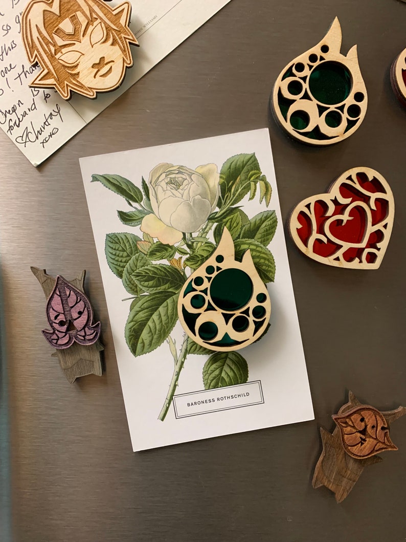 K&ouml;nnte beinhalten: Ein Set aus f&uuml;nf Holzschmuckst&uuml;cken mit filigranen Designs und bunten Glasakzenten. Die Schmuckst&uuml;cke sind in Form eines Herzens, einer Tr&auml;ne und eines Blattes gestaltet. Die Schmuckst&uuml;cke werden auf einem silbernen K&uuml;hlschrank mit einer Postkarte mit einer wei&szlig;en Rose pr&auml;sentiert.