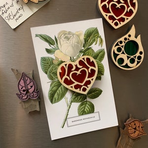 K&ouml;nnte beinhalten: Ein Set aus f&uuml;nf Holz-Laserschnitt-Ornamenten. Die Ornamente haben die Form eines Herzens, einer Blume und eines Blattes. Die Herzornamente sind rot und haben ein detailliertes Design. Das Blumenornament ist wei&szlig; mit gr&uuml;nen Bl&auml;ttern. Das Blatt-Ornament ist gr&uuml;n und hat ein detailliertes Design. Die Ornamente befinden sich auf einem silbernen Hintergrund.