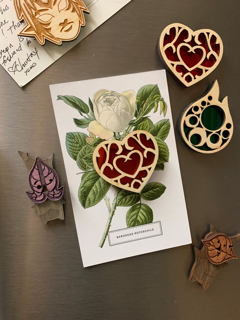 K&ouml;nnte beinhalten: Ein Set aus f&uuml;nf Holz-Laserschnitt-Ornamenten. Die Ornamente haben die Form eines Herzens, einer Blume und eines Blattes. Die Herzornamente sind rot und haben ein detailliertes Design. Das Blumenornament ist wei&szlig; mit gr&uuml;nen Bl&auml;ttern. Das Blatt-Ornament ist gr&uuml;n und hat ein detailliertes Design. Die Ornamente befinden sich auf einem silbernen Hintergrund.