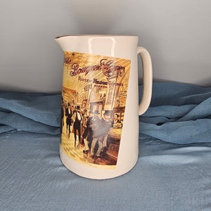 May include: A white ceramic pitcher with a brown handle. The pitcher has a vintage image of a bar scene with the text "Royal Lager" printed on it.