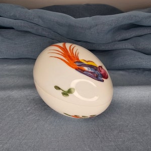 May include: White ceramic egg-shaped trinket box with a painted design of a colorful bird with orange, yellow, red, blue, and green accents.