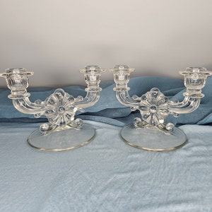 May include: A pair of clear glass candle holders with ornate designs. Each holder has two candle arms and a base with a floral motif.