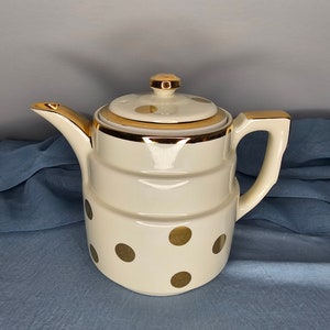May include: A white ceramic teapot with a gold trim and gold polka dots. The teapot has a lid with a small knob on top.