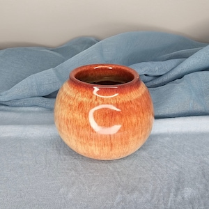 May include: A round ceramic planter with a glossy, orange glaze. The planter has a subtle, striped pattern and a slightly rounded rim.