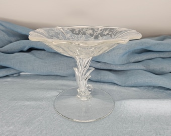 May include: A clear glass pedestal bowl with a floral design. The bowl has a wide, scalloped rim and a delicate stem with a floral motif.