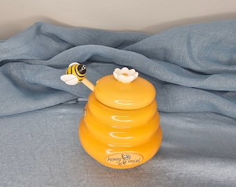 May include: A yellow ceramic honey pot shaped like a beehive with a white flower lid and a wooden bee dipper. The pot has the text "honey & miel" on the bottom.