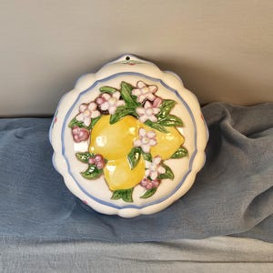 May include: Round ceramic wall hanging featuring a lemon design. The white background is adorned with a blue scalloped edge, yellow lemons, green leaves, and pink and white flowers. A small hole is at the top for hanging.