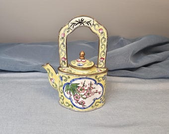 May include: A decorative yellow teapot with a matching lid and a gold handle. The teapot is adorned with floral designs in pink, green, and blue. The handle and trim are gold.