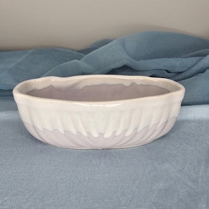Könnte beinhalten: Ein weißer und helllilafarbener Keramik-Pflanztopf mit wellenförmigem Design. Der Topf ist oval geformt und hat eine glatte, glänzende Oberfläche.