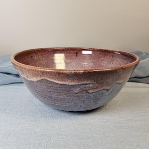 Könnte beinhalten: Eine große, handgefertigte Keramikschüssel mit einer lila, rosa und weißen Glasur. Die Schüssel hat einen breiten, abgerundeten Rand und einen leicht ausgestellten Boden.