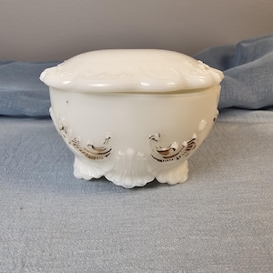 May include: A white, lidded trinket dish with a scalloped rim and base. The dish features gold-colored decorative accents and a rounded shape. The overall design is vintage and elegant, suitable for storing small items.