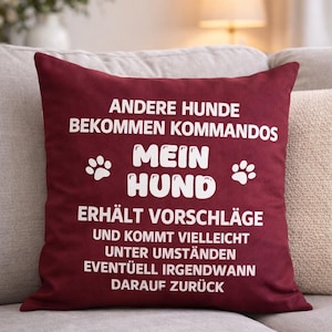 May include: A burgundy throw pillow with white text. The text reads "ANDERE HUNDE BEKOMMEN KOMMANDOS MEIN HUND ERHÄLT VORSCHLÄGE UND KOMMT VIELLEICHT UNTER UMSTÄNDEN EVENTÜELL IRGENDWANN DARAUF ZURÜCK". Paw prints are also included.