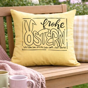 May include: A yellow decorative pillow with a black graphic that says "frohe OSTERN" in a whimsical font, with a bunny outline. The pillow is on a wooden bench with other pillows and mugs.
